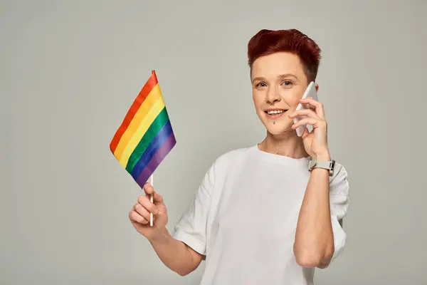 Feliz pelirroja no binaria persona sosteniendo pequeña bandera LGBT y hablando en el teléfono móvil en gris - foto de stock
