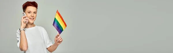 Feliz pelirroja queer persona sosteniendo pequeña bandera LGBT y hablando en el teléfono inteligente en gris, bandera - foto de stock