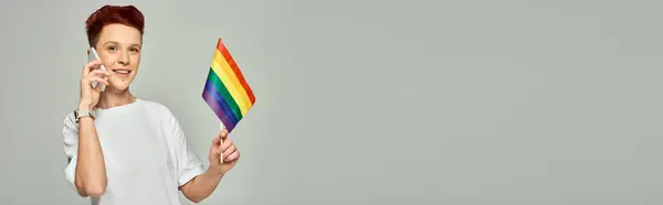 Felice persona queer rossa che tiene piccola bandiera LGBT e parla su smartphone su grigio, banner — Foto stock