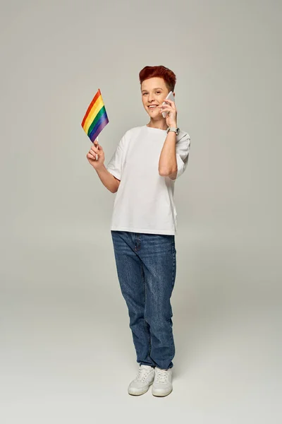 Feliz pelirroja queer persona sosteniendo pequeña bandera LGBT y hablando en el teléfono inteligente en gris, bandera - foto de stock