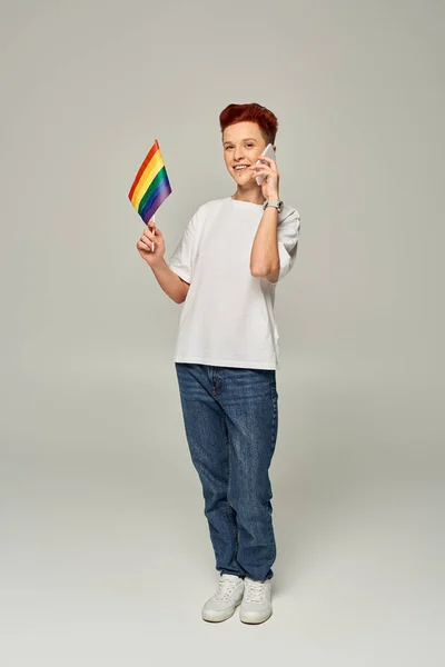 Happy redhead queer person holding small LGBT flag and talking on smartphone on grey, banner — Stock Photo