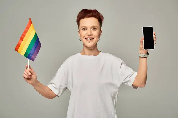 Joyful redhead queer person holding small LGBT flag and smartphone with blank screen on grey — Stock Photo
