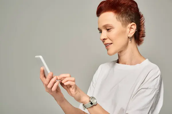 Smiling redhead queer person in white t-shirt messaging on mobile phone while standing on grey — Stock Photo