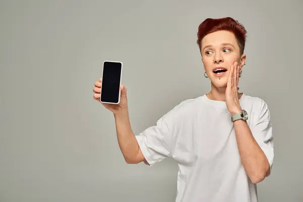 Amazed redhead queer person in white t-shirt showing smartphone with blank screen on grey — Stock Photo