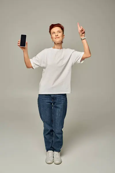 Queer persona sosteniendo smartphone con pantalla en blanco mientras mira hacia arriba y señala con el dedo en gris - foto de stock