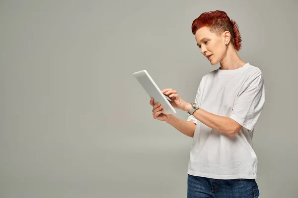 Redhead non-binary person in white t-shirt using digital tablet while standing on grey backdrop — Stock Photo
