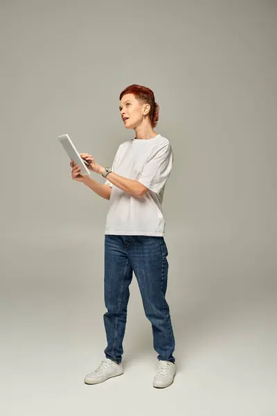 Nachdenkliche nichtbinäre Person in weißem T-Shirt mit digitalem Tablet und Blick weg auf grauem Hintergrund — Stockfoto