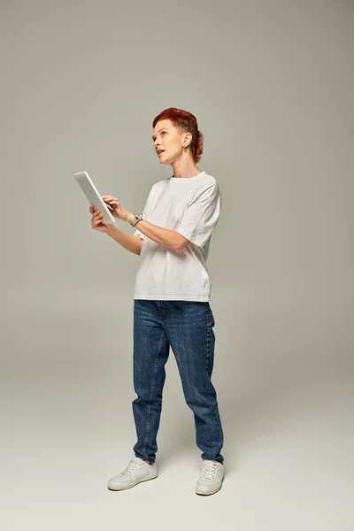 Personne réfléchie non-binaire en t-shirt blanc en utilisant une tablette numérique et en regardant loin sur fond gris — Photo de stock