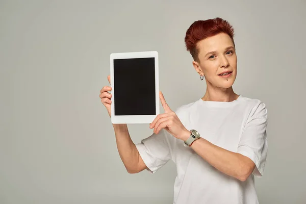 Pelirroja persona no binaria en camiseta blanca mostrando tableta digital con pantalla en blanco sobre fondo gris - foto de stock
