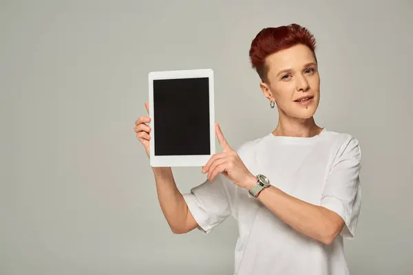 Redhead non-binary person in white t-shirt showing digital tablet with blank screen on grey backdrop — Stock Photo
