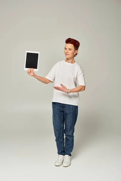 Redhead non-binary person in white t-shirt showing digital tablet with blank screen on grey backdrop — Stock Photo