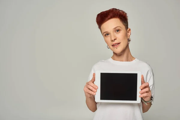Pelirroja queer persona en camiseta blanca mostrando tableta digital con pantalla en blanco sobre fondo gris - foto de stock