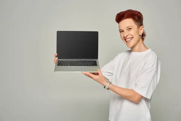 Pelirroja queer persona en blanco camiseta mostrando portátil con pantalla en blanco sobre fondo gris - foto de stock