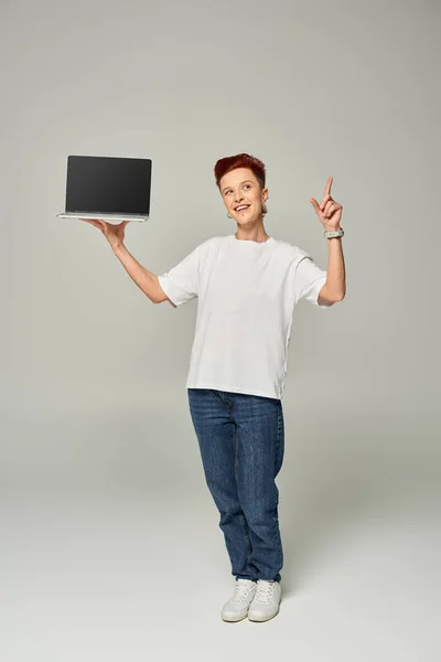 Sonriente freelancer queer mostrando signo de idea mientras sostiene el ordenador portátil con pantalla en blanco sobre fondo gris - foto de stock