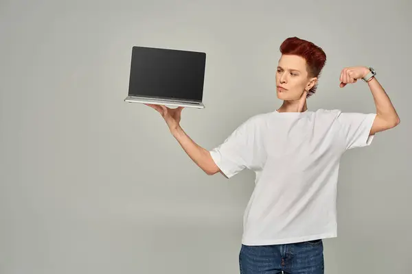 Freelancer queer confiado mostrando músculos mientras sostiene el ordenador portátil con pantalla en blanco sobre fondo gris - foto de stock