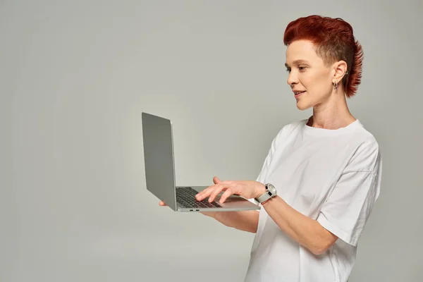 Redhead queer freelancer in white t-shirt networking on laptop while standing on grey backdrop — Stock Photo