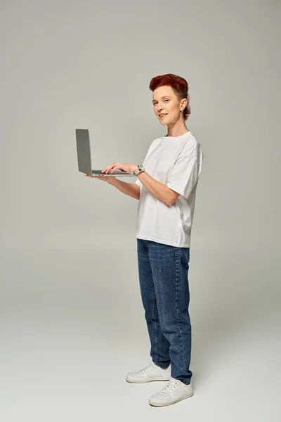 Redhead queer freelancer in white t-shirt standing with laptop and looking at camera on grey — Stock Photo