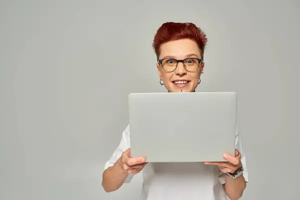 Amazed queer freelancer in white t-shirt and eyeglasses holding laptop and looking at camera on grey — Stock Photo