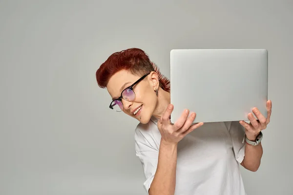 Allegra rossa queer freelance in occhiali che tengono il computer portatile e guardando la fotocamera su grigio — Foto stock