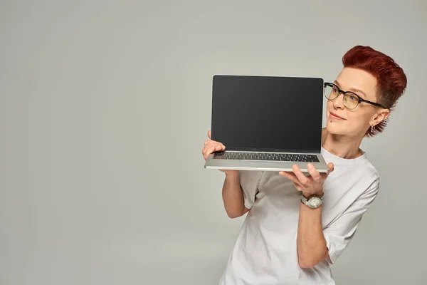 Sonriente pelirroja queer freelancer en gafas portátiles con pantalla en blanco sobre fondo gris - foto de stock