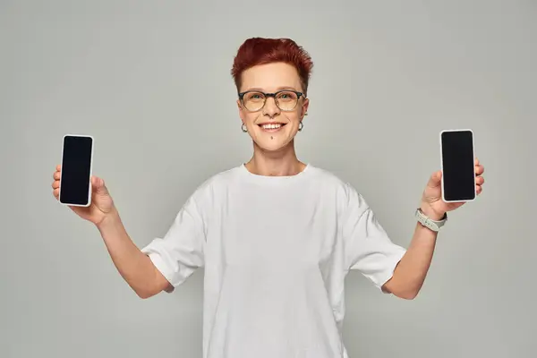 Pelirroja sonriente queer persona en gafas que muestra teléfonos inteligentes con pantalla en blanco sobre fondo gris - foto de stock