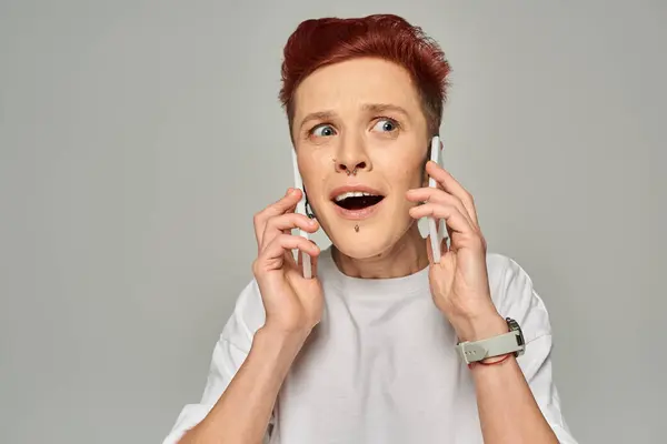 Beeindruckt und erstaunt rothaarige queere Person mit offenem Mund, die auf Smartphones vor grauem Hintergrund spricht — Stockfoto