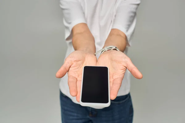 Vue recadrée de la personne tenant un téléphone portable avec écran blanc sur fond gris, smartphone dans les mains — Photo de stock