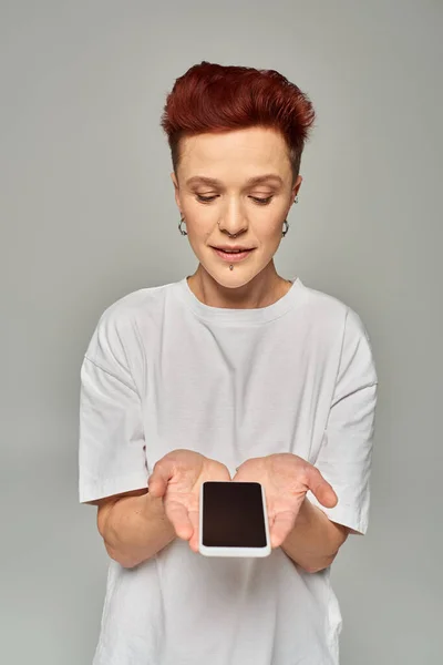 Redhead non-binary person in white t-shirt holding mobile phone with blank screen on grey backdrop — Stock Photo