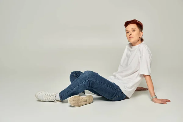 Elegante rossa non binaria persona in bianco t-shirt e jeans seduto e guardando la fotocamera sul grigio — Foto stock
