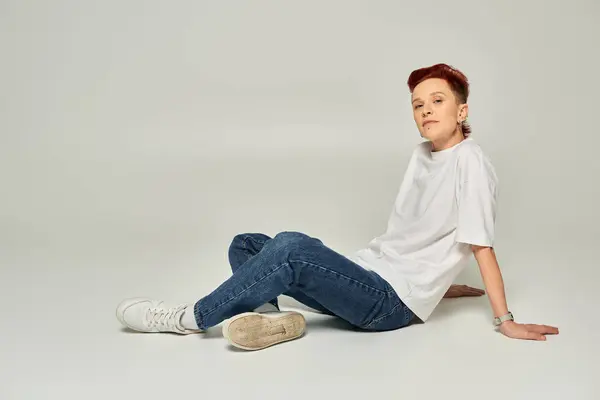 Elegante pelirroja persona no binaria en camiseta blanca y jeans sentado y mirando a la cámara en gris - foto de stock