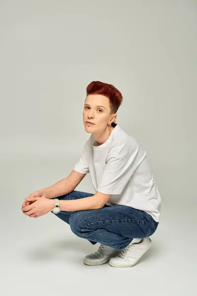 Rothaarige, queere Person in weißem T-Shirt und Jeans, die auf den Hinterbeinen sitzt und in die Kamera auf grau blickt — Stockfoto