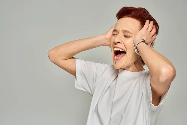 Personne queer émotionnelle en t-shirt blanc touchant la tête et criant avec les yeux fermés sur gris — Photo de stock