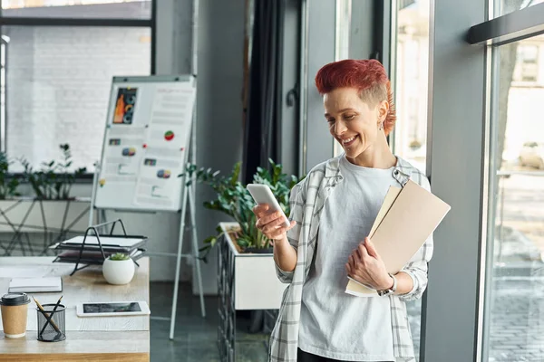 Щаслива квір-людина в повсякденному вбранні, що стоїть з документами в офісі та повідомленнями на смартфоні — стокове фото