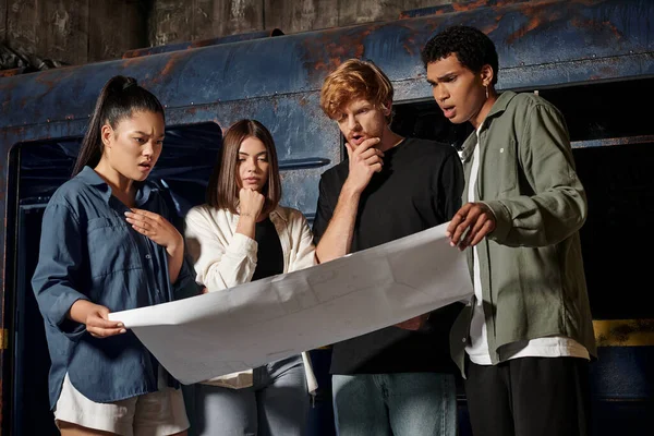 Grupo de amigos pensativos jóvenes multiculturales que resuelven la tarea de búsqueda mientras miran el mapa en la sala de escape - foto de stock
