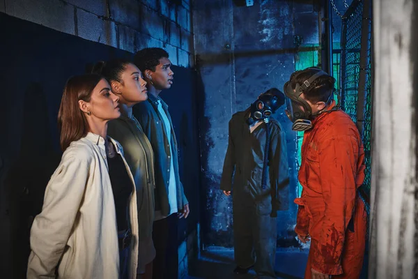 Personas con máscaras de gas de pie cerca de amigos interracial aterrorizados durante la búsqueda en la sala de escape, peligro - foto de stock