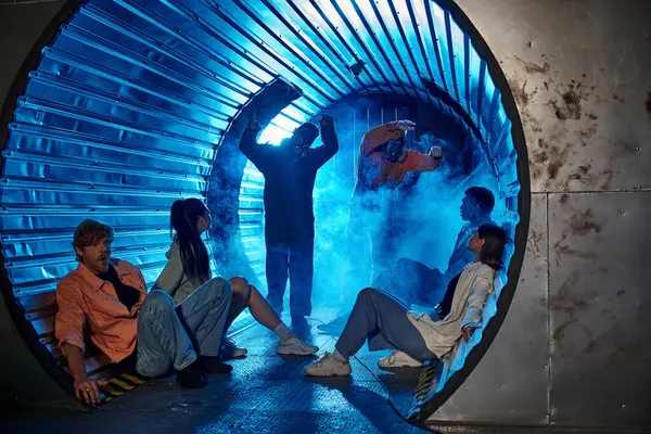 Amigos multiétnicos sentados en un túnel cerca de gente rara con máscaras de gas, aventura en la sala de misiones - foto de stock