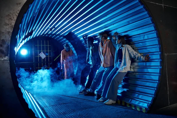 Amigos multiétnicos em pé no túnel perto de pessoas bizarras em máscaras de gás, aventura sala de busca — Fotografia de Stock