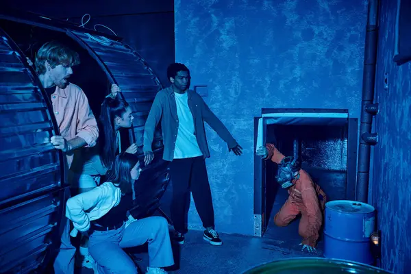 Мультикультурная группа исследователей, скрывающихся от человека в противогазе во время побега из комнаты квестов — стоковое фото