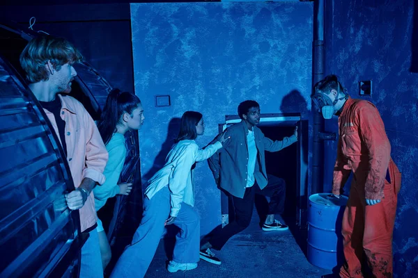 Grupo multicultural de amigos escapando del hombre con máscara de gas durante la aventura en la sala de misiones - foto de stock