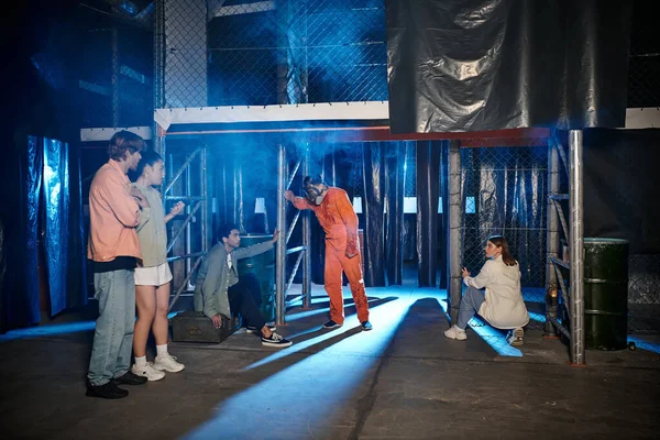 Person in orangefarbenem Anzug und Gasmaske neben Gruppe verängstigter Freunde im Escape Room — Stockfoto
