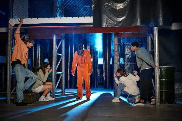 Pessoa assustadora em terno laranja ppe e máscara de gás perto de jovens amigos inter-raciais na sala de fuga — Fotografia de Stock