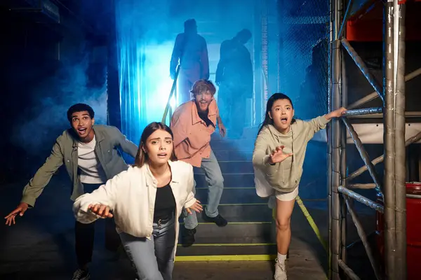 Vier junge multikulturelle Freunde laufen in Gasmasken und Schutzanzügen vor furchterregenden Menschen im Escape Room davon — Stockfoto