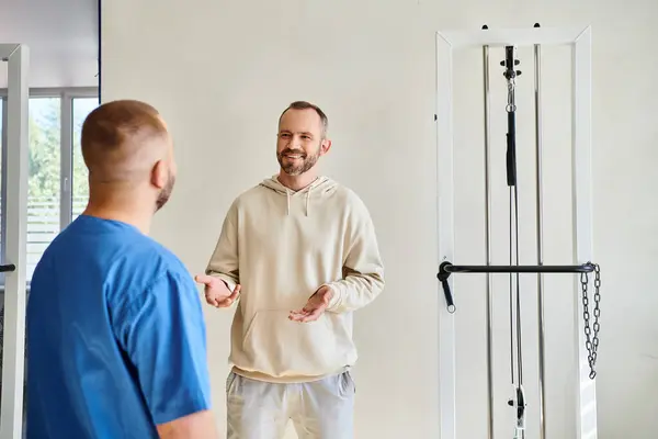 Uomo positivo che parla con il medico in uniforme blu durante l'appuntamento nel centro di riabilitazione kinesio — Foto stock