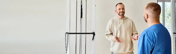 Усміхнений чоловік розмовляє з фахівцем у блакитній формі під час зустрічі в центрі кінезіо, банер — стокове фото