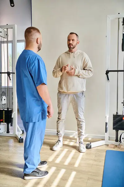 Bell'uomo che discute il piano di trattamento con il medico in uniforme blu nel centro di recupero kinesio — Foto stock