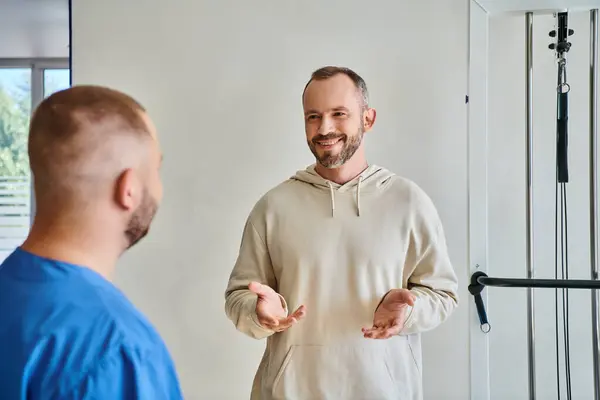 Счастливый бородатый мужчина жестикулирует и разговаривает с молодым специалистом реабилитационного центра кинезиотерапии — стоковое фото