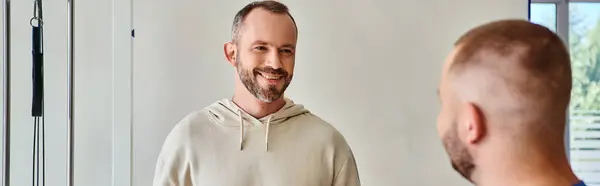 Hombre barbudo guapo sonriendo cerca de joven especialista en recuperación en el centro kinesio, pancarta horizontal - foto de stock