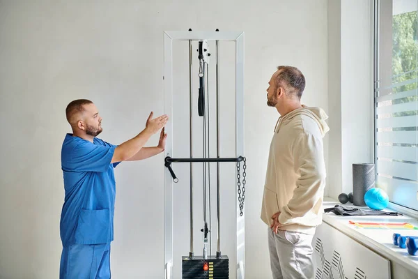 Réhabilitologue en uniforme bleu montrant l'équipement d'exercice à son patient dans le centre de kinesio — Photo de stock