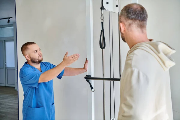 Молодий лікар у блакитній формі показує тренажер для людини в реабілітаційному центрі Кінезіо — стокове фото