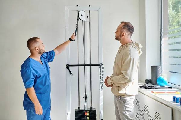 Réhabilitologue professionnel en uniforme bleu montrant la machine de formation à l'homme dans le centre de kinésiologie — Photo de stock